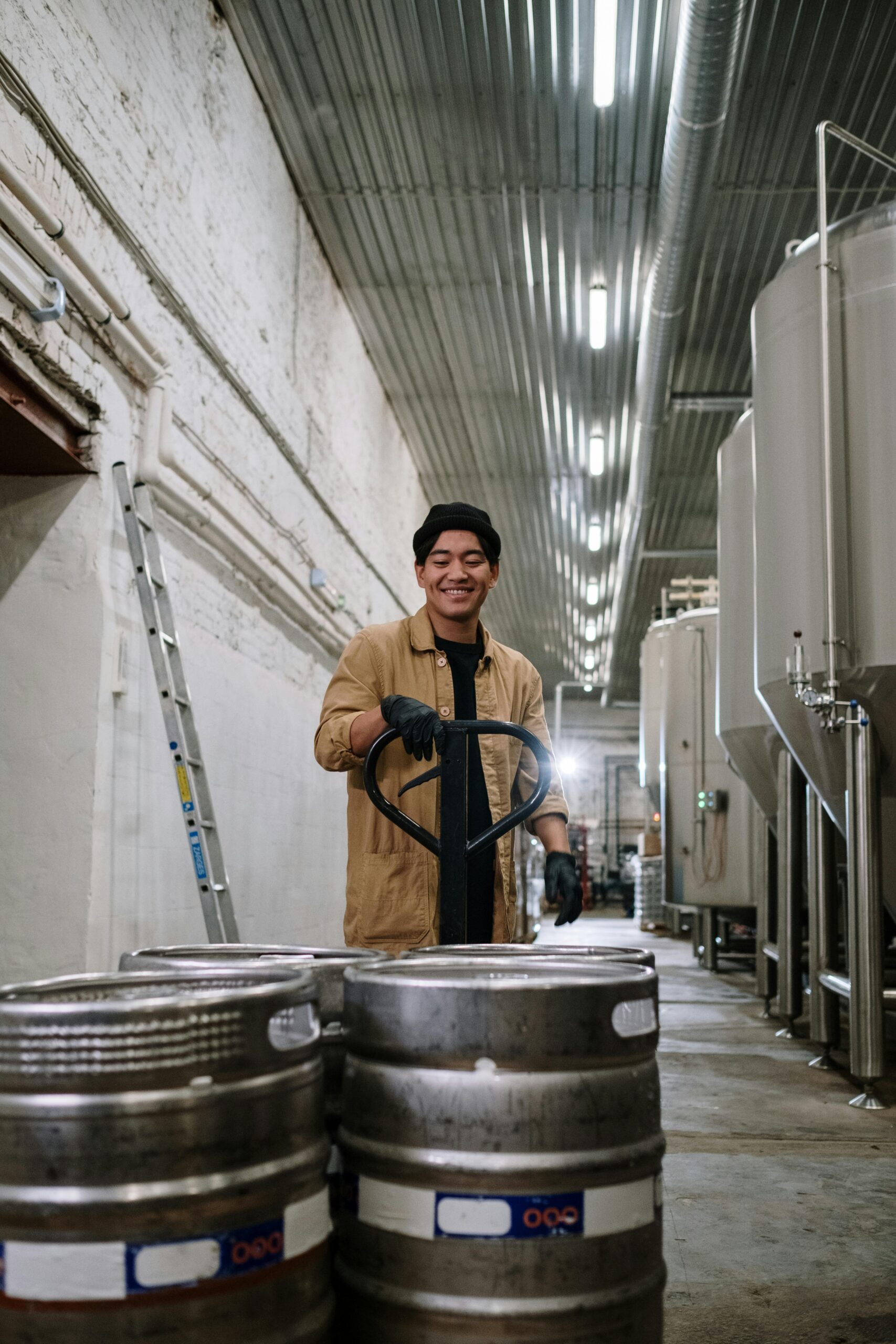 Man smiling while pulling beer kegs inside a brewery, showcasing industrial production.